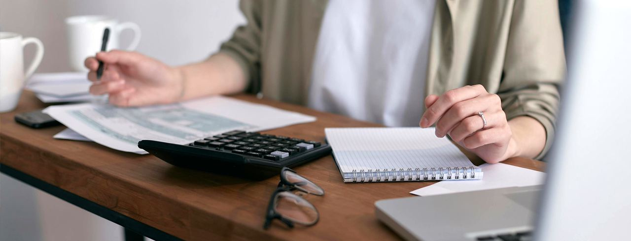 a closeup of a desk with papers, a calculator, glasses, and hands holding a pen