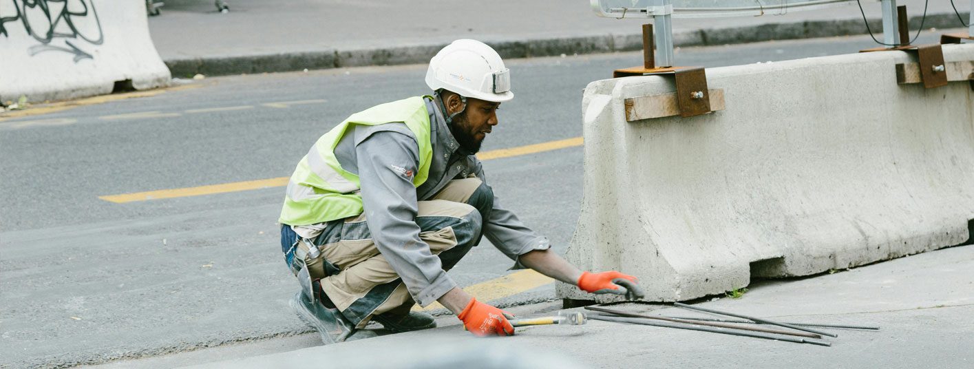 a man in a safety vest and construction hat squatting in the middle of a road next to a concrete barrier